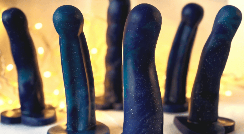 6 'starry night' ( dark blue marbled with purple) limited edition Sh! silicone dildos astanding upright against a warm, softly blurred background with out-of-focus lights.