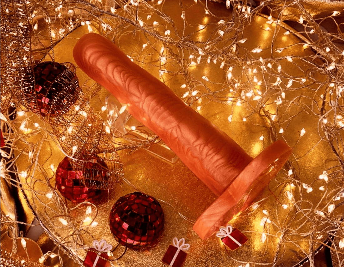 A gold silicone dildo displayed on a golden surface surrounded by warm fairy lights, red Christmas ornaments, and small gift boxes.